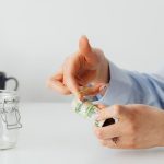 Close-up of hands rolling US dollars with a rubber band near an empty glass jar.